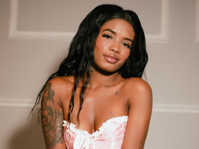 LillieBurges with dark wavy hair, tan skin, and tattoos on her right arm, wearing a pink, ruffled, strapless top, pouting slightly with full, glossy lips.