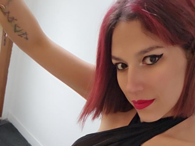 LilyDiabless with red-dyed hair, black eyeliner, red lipstick, and a black top, showing her tattooed arm and a sexy smile. Boobs partially visible, arm raised.