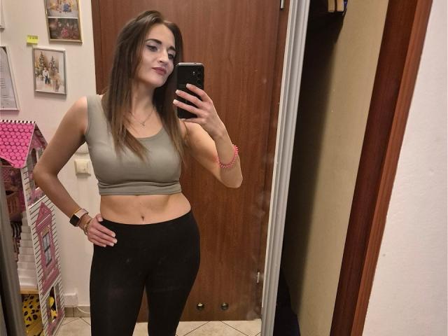 NicoletteCreazy with long brown hair, wearing a gray sports bra and black leggings, taking a mirror selfie, showing her toned midriff and small ass. She's smiling slightly.