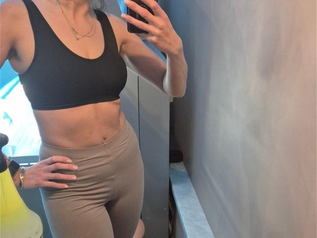 NicoletteCreazy with a slim, toned body, wearing a black sports bra and tight beige leggings, showing her flat stomach and small ass, taking a mirror selfie.
