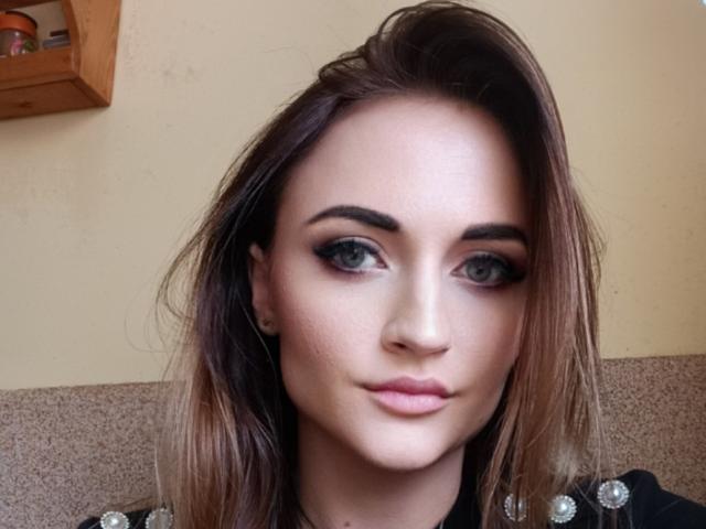 NicoletteCreazy has a cute face with blue eyes, thick dark eyebrows, and pink lipstick. She's got long brown hair and a slight smile, looking straight at the camera.