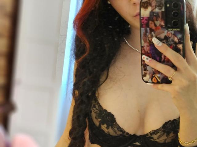 LucyWilow with long black and red curly hair, wearing a black lace bra, showing her tits in a mirror selfie, holding a phone with a colorful case.