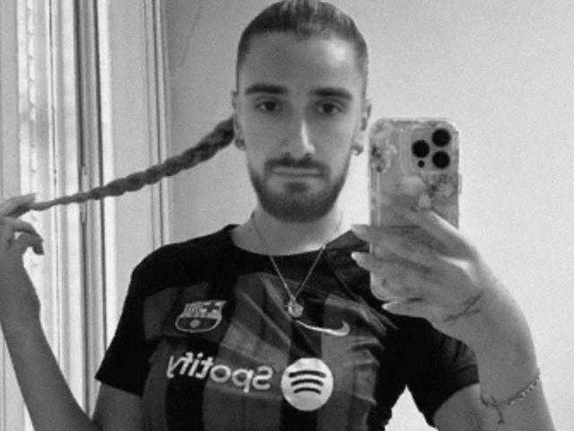 JodyTs with a long braid, holding phone in right hand, wearing black and gray striped soccer shirt, looking at mirror, holding braid with left hand, serious expression.