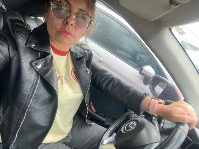 MiaAmoretti with puffy lips, wearing glasses, black leather jacket, and red shirt, sitting in a car, holding the steering wheel with a pink watch.