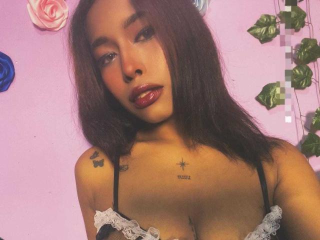 LilithSanz with brown hair, glossy red lips, black lace bra, tattoos on shoulder and chest, and medium tits, against pink wall with rose and vine decorations.