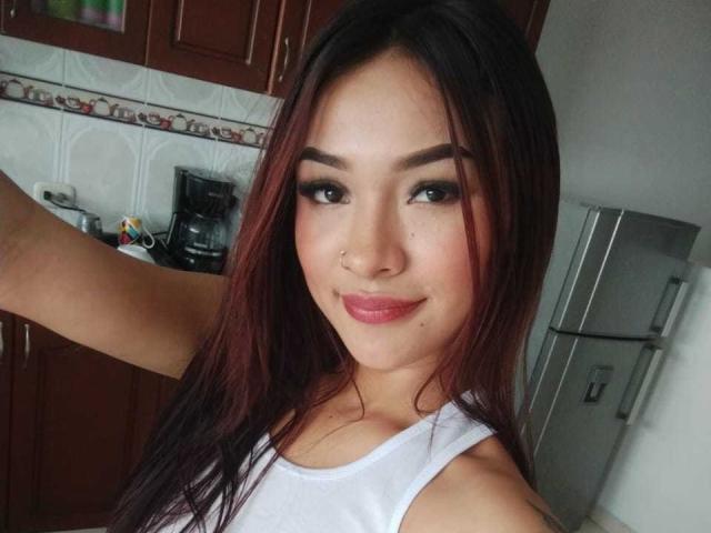 SashaHans with dark brown hair, red highlights, white tank top, nose ring, and pink lipstick, smiling at the camera, showing her tits slightly in the frame.