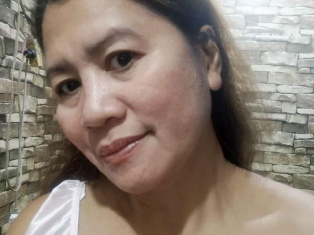 Hannahh69 has a cute smile with slightly flushed cheeks, brown eyes, and light brown hair. She's wearing a white strap, showing her bare shoulders and soft skin.