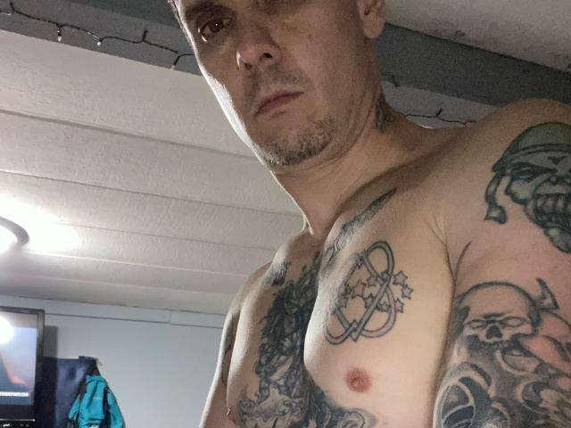 Cermisyy, shirtless, with tattoos on chest and arms, bald with light beard, small nipples, slightly muscular, looking down at camera, in dimly lit room.