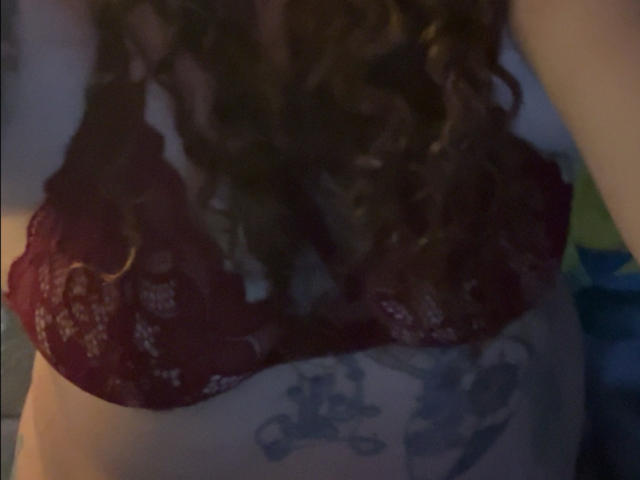 Cermisyy's red lace bra, showing her boobs, with curly brown hair, and a tattoo of a motorcycle below her tits. Dim, dark room.