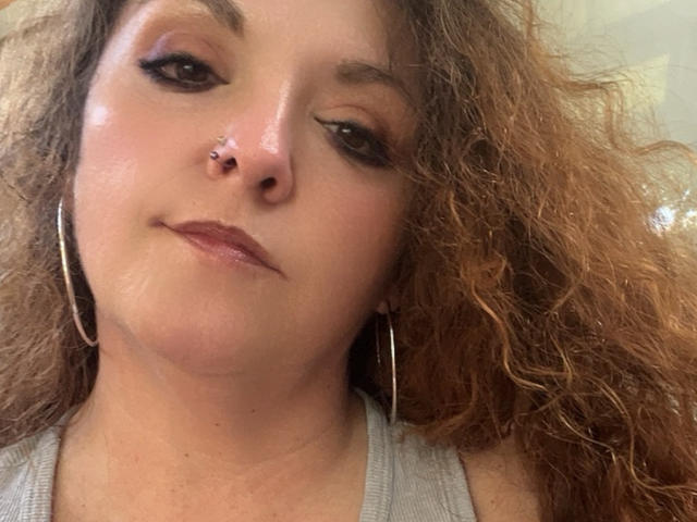 Cermisyy has curly brown hair, a nose piercing, black eyeliner, and is wearing hoop earrings with a subtle smile, wearing a gray top. Her skin is slightly shiny.