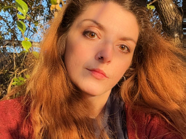 Cermisyy has long, wavy, reddish-brown hair, brown eyes, a small nose ring, and pink lips. She wears a red shirt, looking slightly to the side.