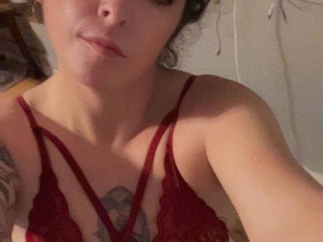 Cermisyy with pale skin, dark curly hair, and red lace bra, showing her small tits and a tattoo under her chest, smiling slightly at the camera.