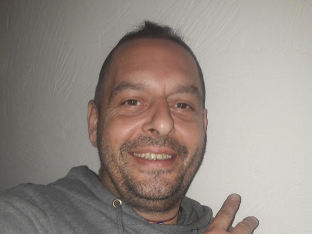 EtnikSatyr hat einen glatzköpfigen Kopf, hellbraune Haut und einen Bart mit einem kleinen Schnurrbart, lächelt mit freiliegenden Zähnen, trägt eine graue Kapuzenjacke und macht eine Rocker-Geste..