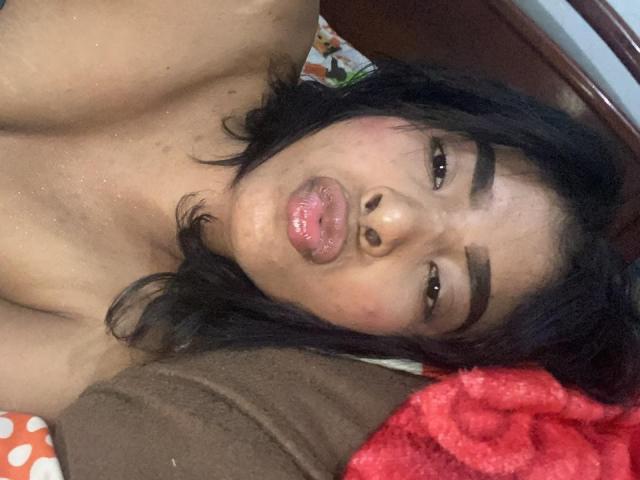 SweetGirland with pouty pink lips, dark wavy hair, and thick eyebrows, lying topless on a bed, looking up with a slightly annoyed or confused expression.