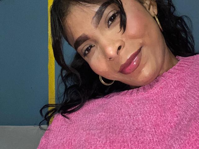 SweetGirland has glossy pink lips, dark wavy hair, thick eyebrows, and wears a pink sweater with gold hoop earrings, smiling slightly. Her face is slightly tilted to the right.