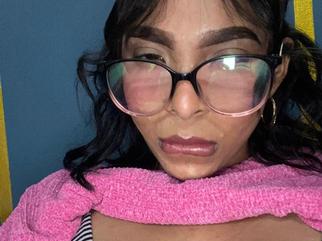 SweetGirland with dark hair, glasses, and pink towel around neck, has wet lipstick and a glistening face, looking slightly confused. Her tits are partially visible under the towel.