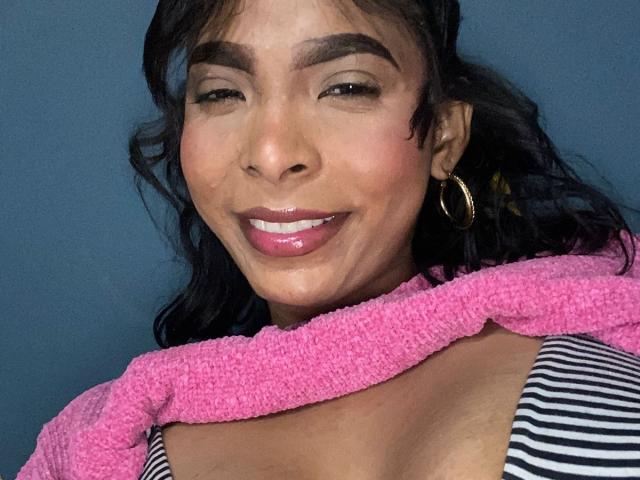 SweetGirland has dark brown skin, black wavy hair, and thick eyebrows. She's smiling with pink glossy lips, wearing a striped top and pink scarf, and gold hoop earrings.