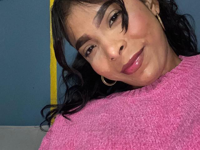 SweetGirland with shiny pink lips, dark wavy hair, and hoop earrings, wearing a pink sweater, smiling slightly, with a yellow pole in the background.