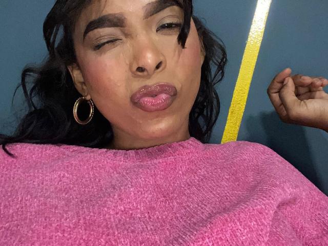 SweetGirland with dark hair, gold hoop earrings, pink sweater, puckering glossy pink lips, winking, and showing a small bruise on her cheek. Her right hand is raised.