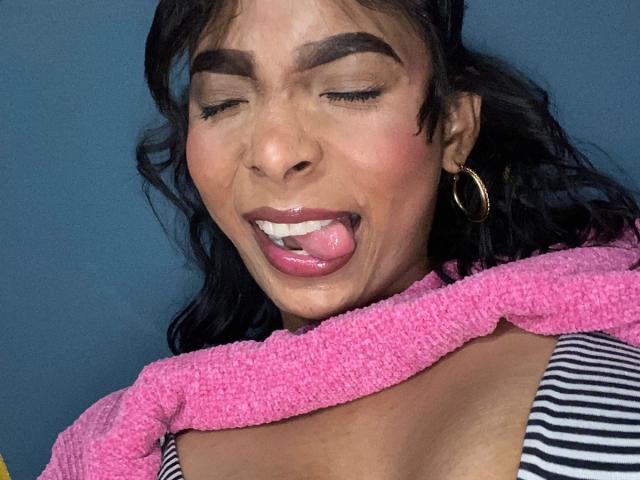 SweetGirland has dark brown hair, thick eyebrows, and is sticking out her tongue with a tooth gap, wearing a pink towel and striped top, with hoop earrings.