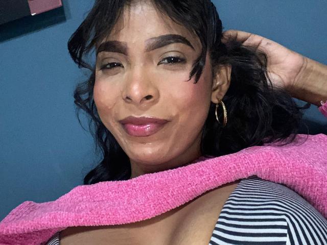 SweetGirland has dark brown hair, thick eyebrows, pink lipstick, gold hoop earrings, and a pink sweater covering her striped top and showing her big, shiny tits.