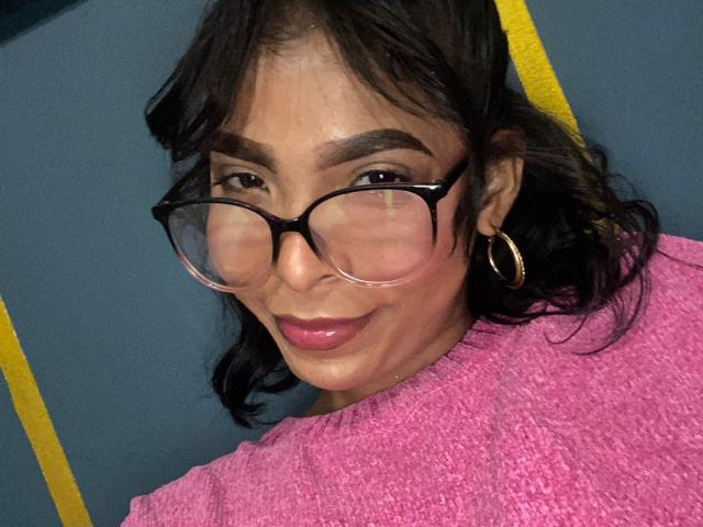 SweetGirland with black glasses, pink lipstick, and pink sweater, smiling, showing her face and hoop earrings, dark wavy hair, and a slightly tan complexion.