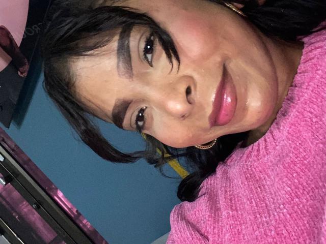 SweetGirland's face with dark, wavy hair, thick eyebrows, shiny pink lips, and a subtle smile, wearing a pink textured shirt, looking slightly to the side.