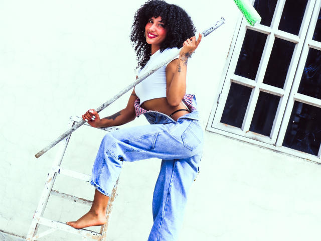 Cinthia Walker, avec ses cheveux noirs bouclés, portant un débardeur blanc, un jean bleu et pieds nus, tenant un balai sur une échelle, souriant avec des lèvres rouges, montrant des tatouages sur son bras..
