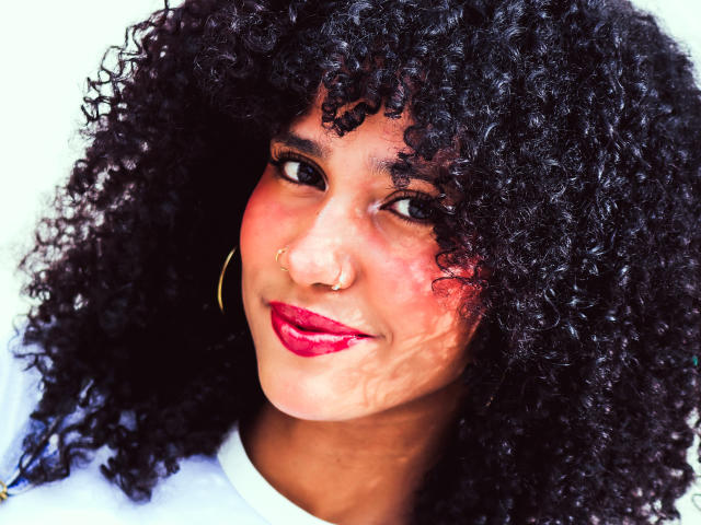 Cinthia Walker, con i capelli neri ricci, rossetto rosso, orecchini a cerchio, piercing al naso e un sorriso sicuro. La sua pelle marrone scura ha un leggero rossore sulle guance..