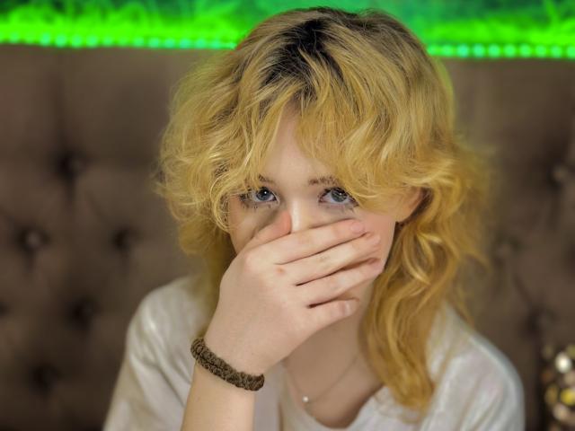 EmilyAngels with blonde, wavy hair, blue eyes, and a white shirt, covering her mouth with her hand, wearing a brown beaded bracelet, looking nervous or shy.