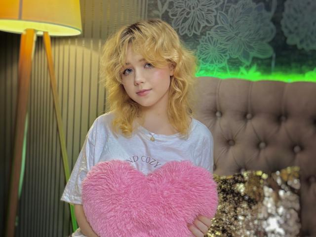 EmilyAngels with curly blonde hair, white tee, holding pink fuzzy heart, standing near gold lamp, tufted beige headboard, and green light. Her small tits are slightly visible.