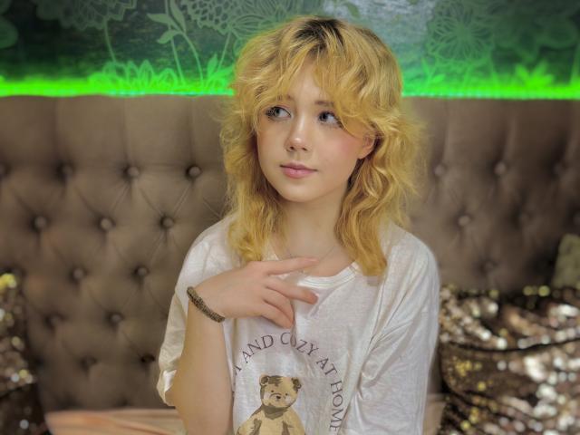 EmilyAngels with blonde wavy hair, wearing a white tee, standing with one hand near her chest, looking to the side, in a tufted brown headboard bedroom.