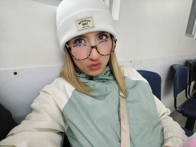 Kaiix with pouty lips, wearing a white beanie, glasses, and a green and white jacket, has blonde hair and a strap across her chest, sitting in a chair.