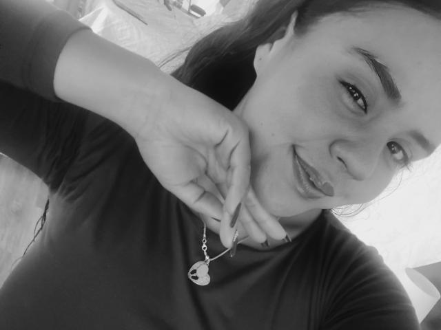 ArielleWilde, with dark hair and a cute smile, holds a necklace with a crescent moon pendant, wearing a dark shirt, in a black-and-white selfie.