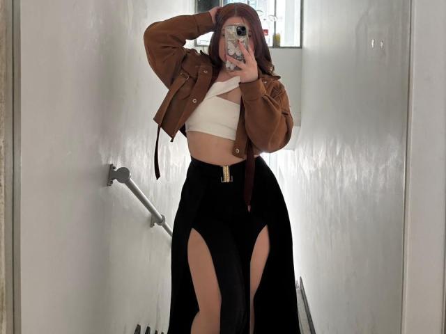 VicluX with long red hair, brown jacket, white crop top, black high-waisted skirt with thigh slits, holding phone, mirror selfie, showing her tits and ass.
