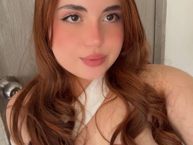 VicluX has long, wavy brown hair, fair skin, and is topless with white fabric covering her tits, showing a slight smile with pink lipstick and black eyeliner.