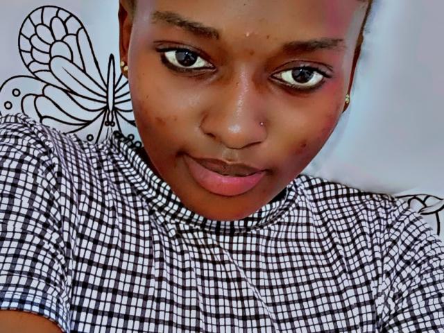 HotVenuss has dark brown skin, wide eyes, small nose, and a subtle smile. She's wearing a black-and-white checkered shirt, with a butterfly tattoo on the left side.