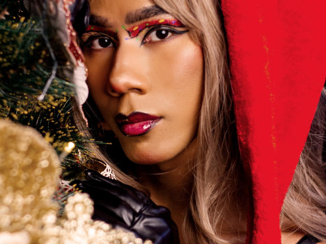 ScarlettHotSweet, con un brillo de labios rosa, un gorro navideño rojo y delineador de ojos de colores, mira fijamente un adorno navideño brillante. Su piel morena y su cabello rubio largo están resaltados..