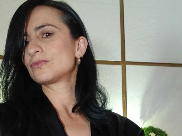 BrendaCantorel has black hair, light skin, small earrings, and is wearing a black shirt. She's slightly pouting with a subtle smile. Her tits are not visible.