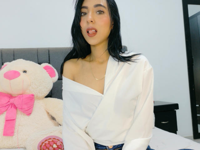 DaphneSpencer has long black hair and fair skin, with her mouth slightly open, revealing a pink tongue. She wears a white, off-shoulder shirt with a subtle sheen, exposing her collarbone and a thin gold necklace. She sits on a bed next to a large, beige teddy bear with pink ears, nose, and a pink bow. The teddy bear has a red heart on its belly. The image has a casual, modern style with soft lighting. DaphneSpencer's expression is relaxed and slightly playful.