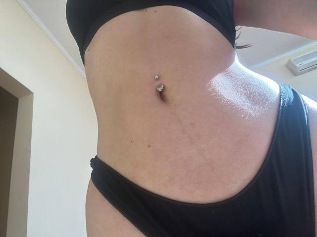 A barriga brilhante e bronzeada de MoonLiza, com um piercing no umbigo, vestindo roupa íntima preta e um sutiã preto, mostrando uma leve curvatura e uma textura de pele lisa..