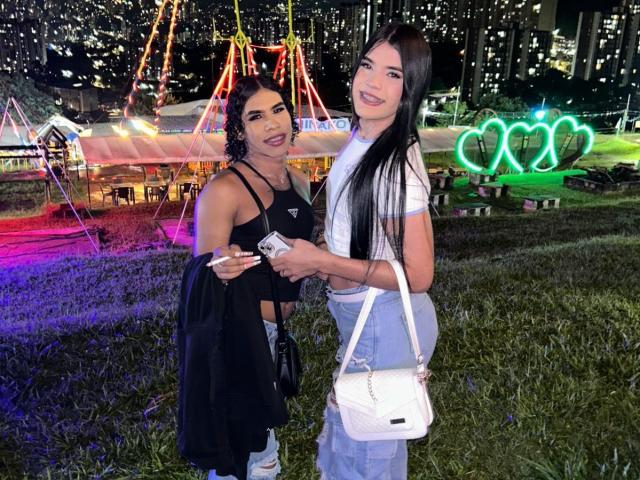 SabrinaAndValery with long black hair, one in a black dress holding a jacket, the other in a white shirt and blue jeans, standing on grass, holding a phone.