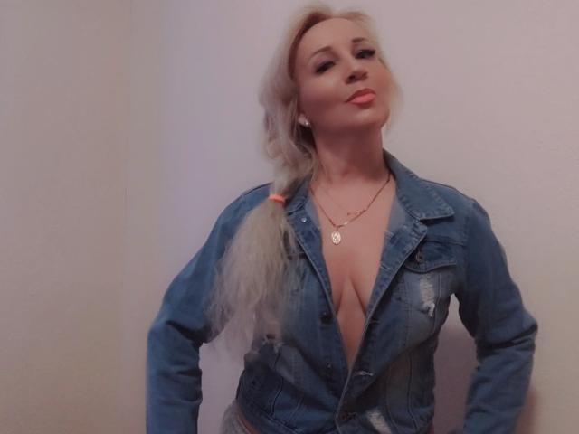 AliceHottie6 is a blonde woman with a long ponytail, wearing a blue denim jacket with a deep V-neck, showing cleavage. She has a slight smile, with pink lipstick and subtle eye makeup. She wears a small necklace with a heart-shaped pendant. Her hands rest on her hips, and her jacket has a few worn patches. The image is slightly blurry. Her skin is fair, and she has a confident, relaxed posture. The overall composition is centered on her upper body.