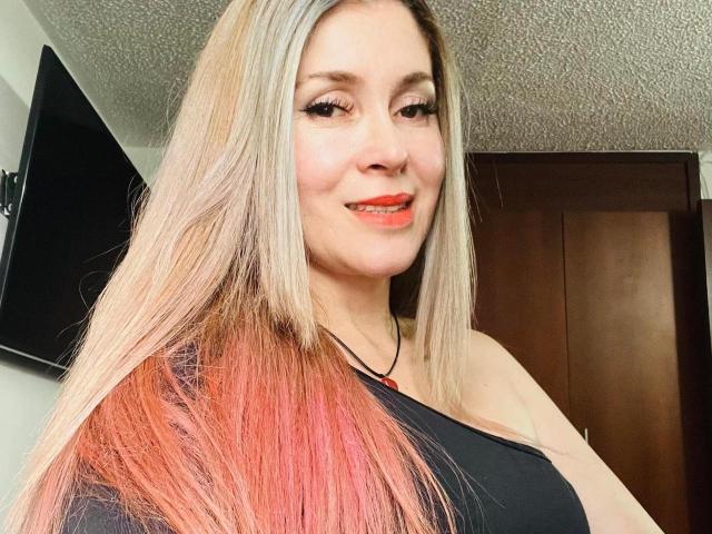 NathalyRobinson with long blonde and pink ombre hair, black top, smiling with red lipstick, and black necklace, showing her tits. She has light skin and a cute face.