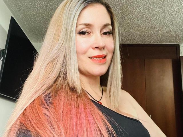 NathalyRobinson has long blonde and pink ombre hair, smiling with red lipstick, wearing a black top, and has a nose ring. Her tits are small and she's cute.