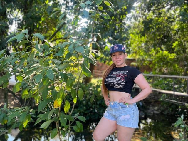 NathalyRobinson with long brown hair, wearing a black crop top, blue denim shorts, and a navy baseball cap, standing in front of a leafy tree. Her tits are small.