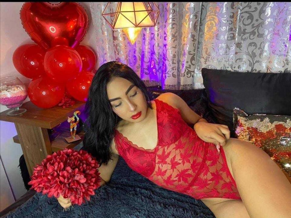 MeganBellx with black hair, red lace lingerie, and red lipstick, holding a red flower bouquet, lying on a black couch, surrounded by red heart balloons and a lamp.