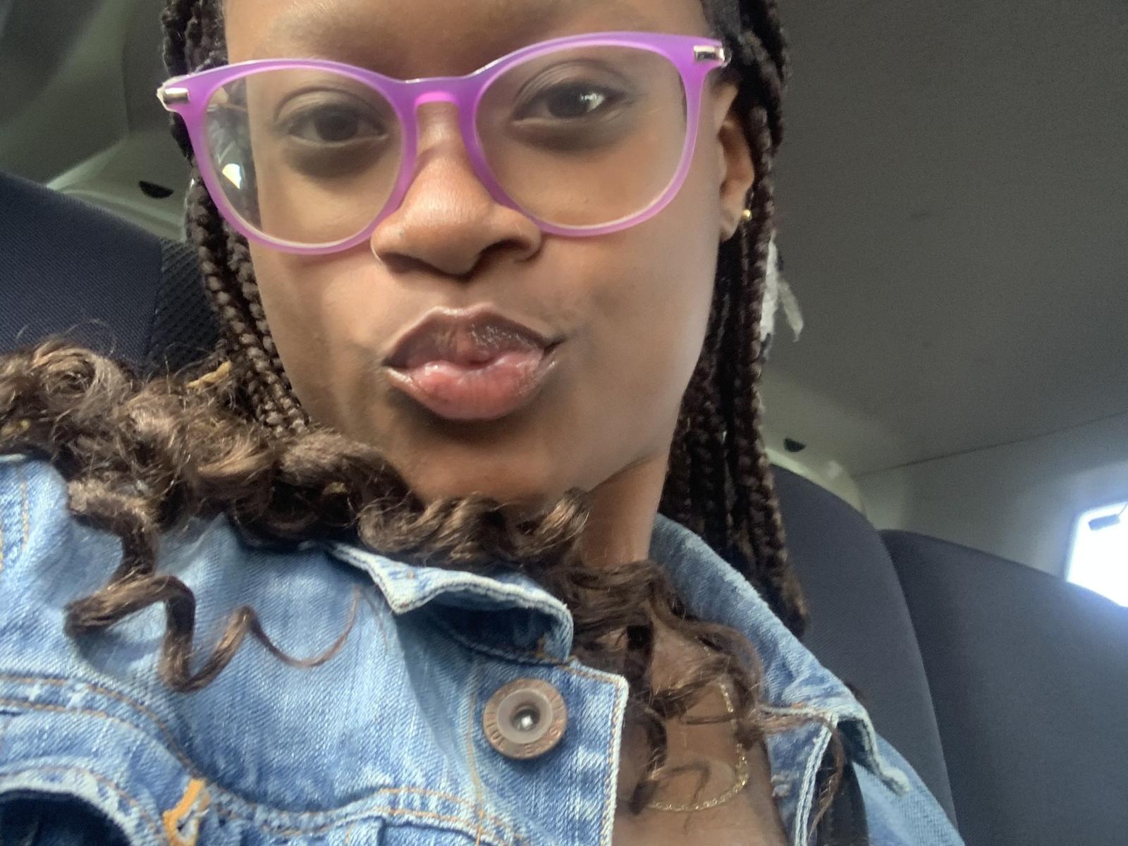 Mssexyy with purple glasses, pouty lips, and curly braids, wearing a blue denim jacket, car interior, black hair, and a gold necklace, looking into the camera.