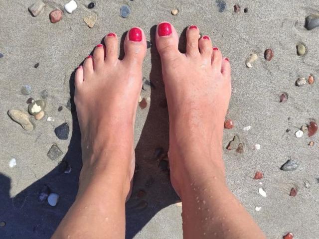 Nicoleta56-hot's feet with red-painted toenails, slightly sandy, standing on a beach with small, colorful pebbles scattered in the sand.