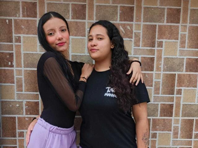 AmarandSofty with black hair, sheer sleeves, and purple pants stands next to AmarandSofty with curly black hair, black shirt, and tattooed arm, both smiling.